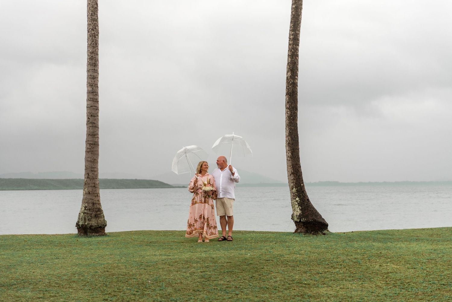 Couple standing between two tall palm trees holding clear umbrellas near a calm tropical waterfront.