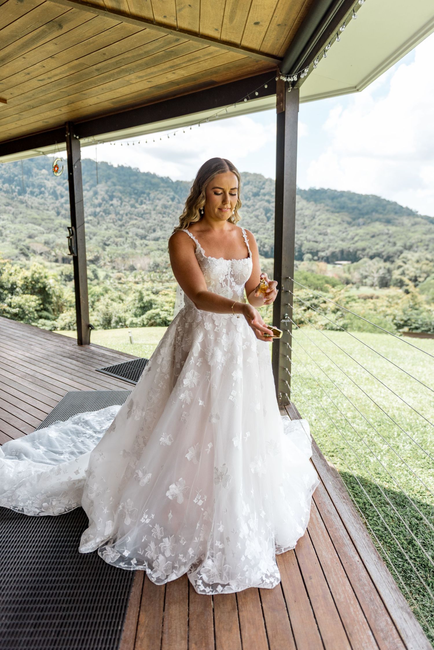 Bride in floral lace wedding gown with long train stands on wooden deck overlooking lush green hills.