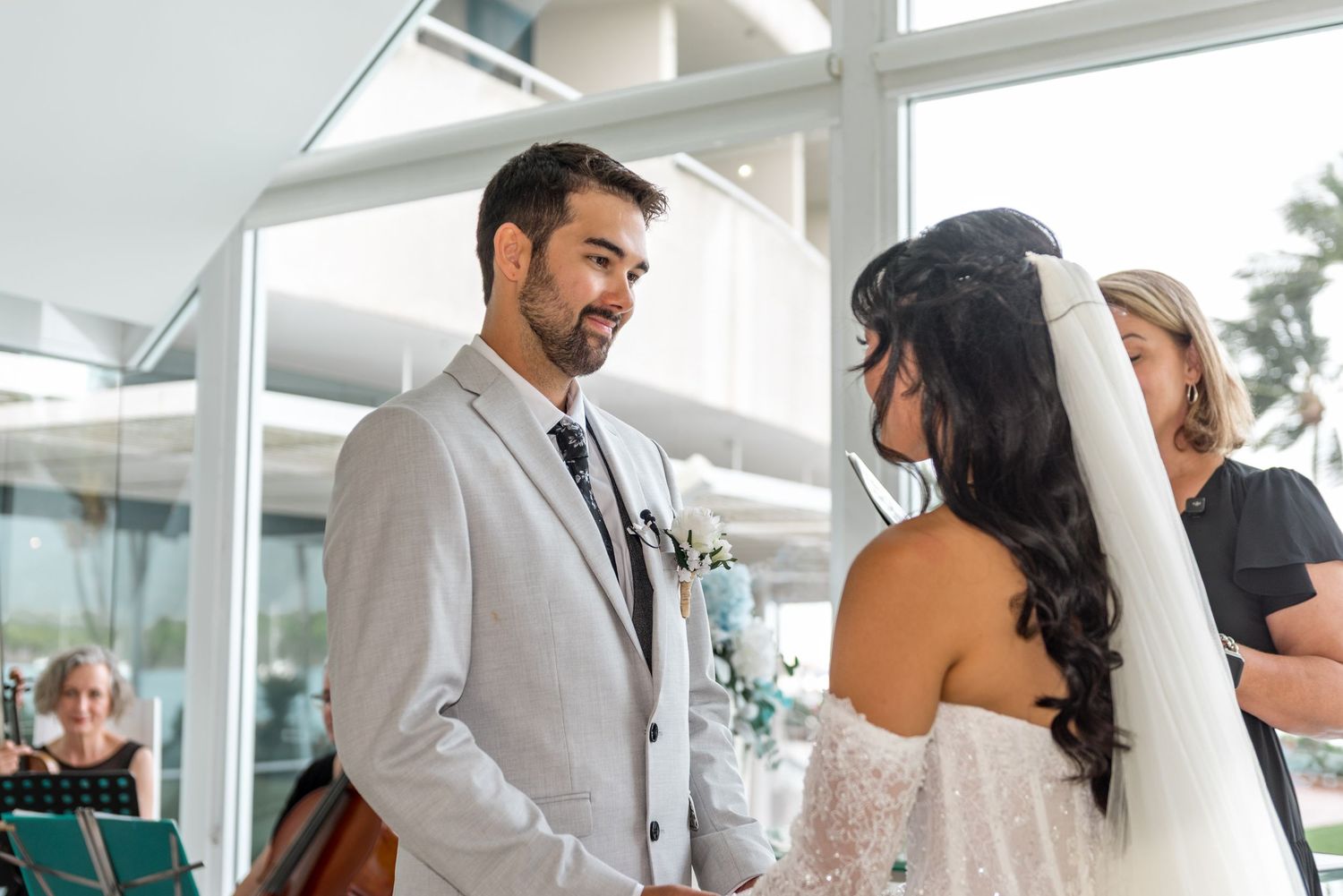 Groom in gray suit smiling at bride during wedding ceremony with officiant in background.