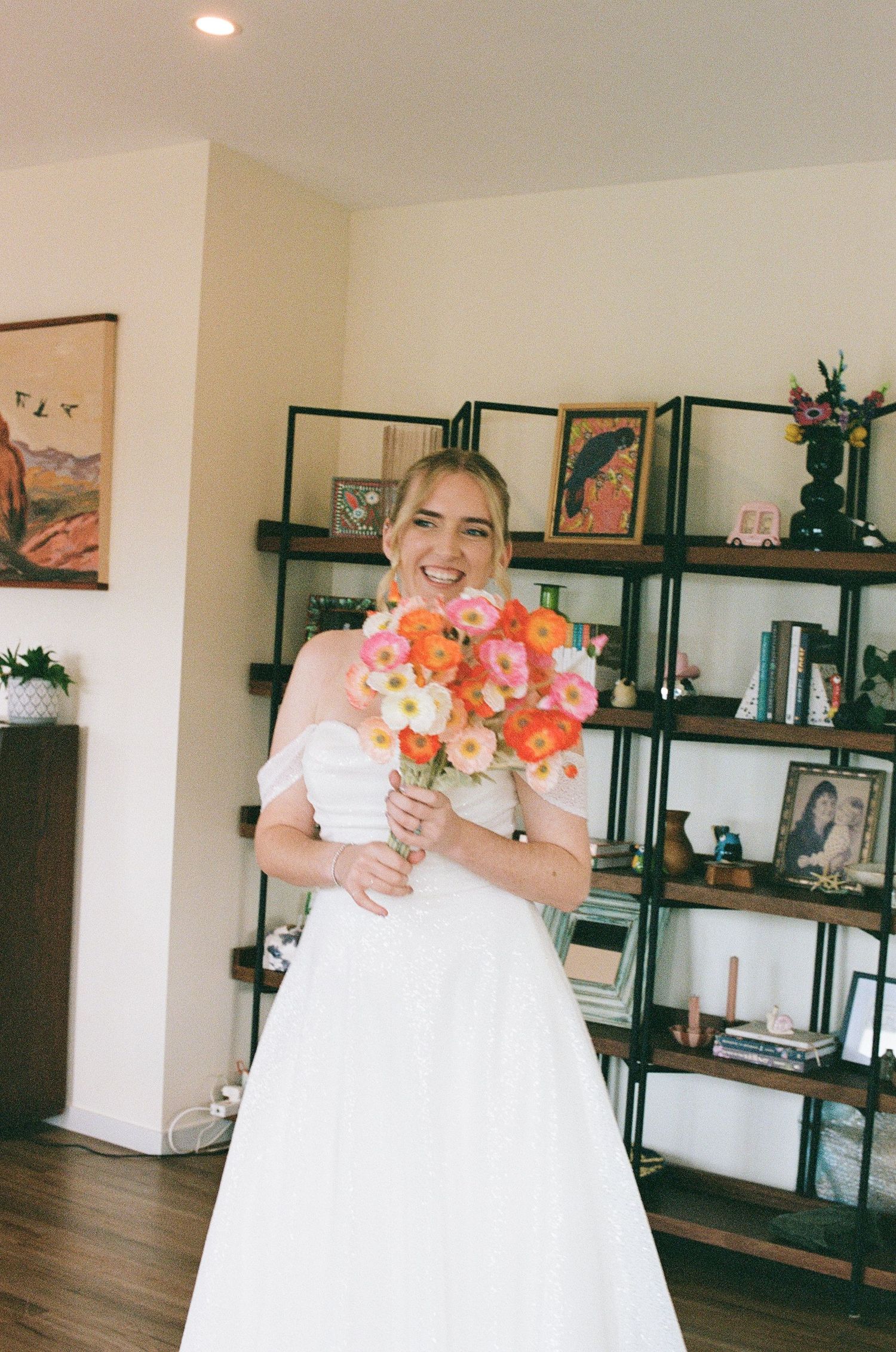 Smiling bride in white off-shoulder gown holding colorful pink and orange bouquet indoors.