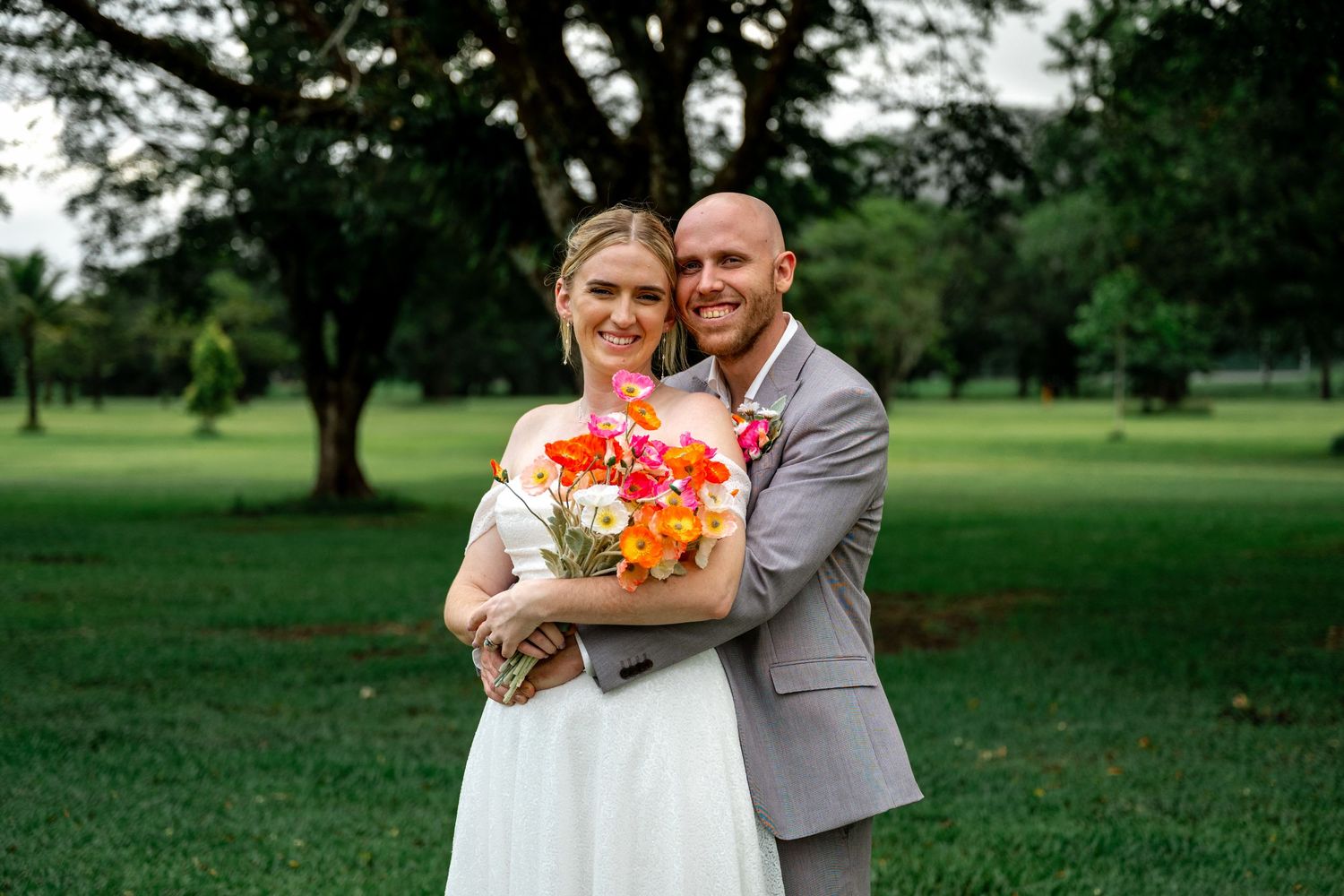 Smiling bride and groom embracing outdoors, bride holding bright orange and pink floral bouquet in lush garden setting.