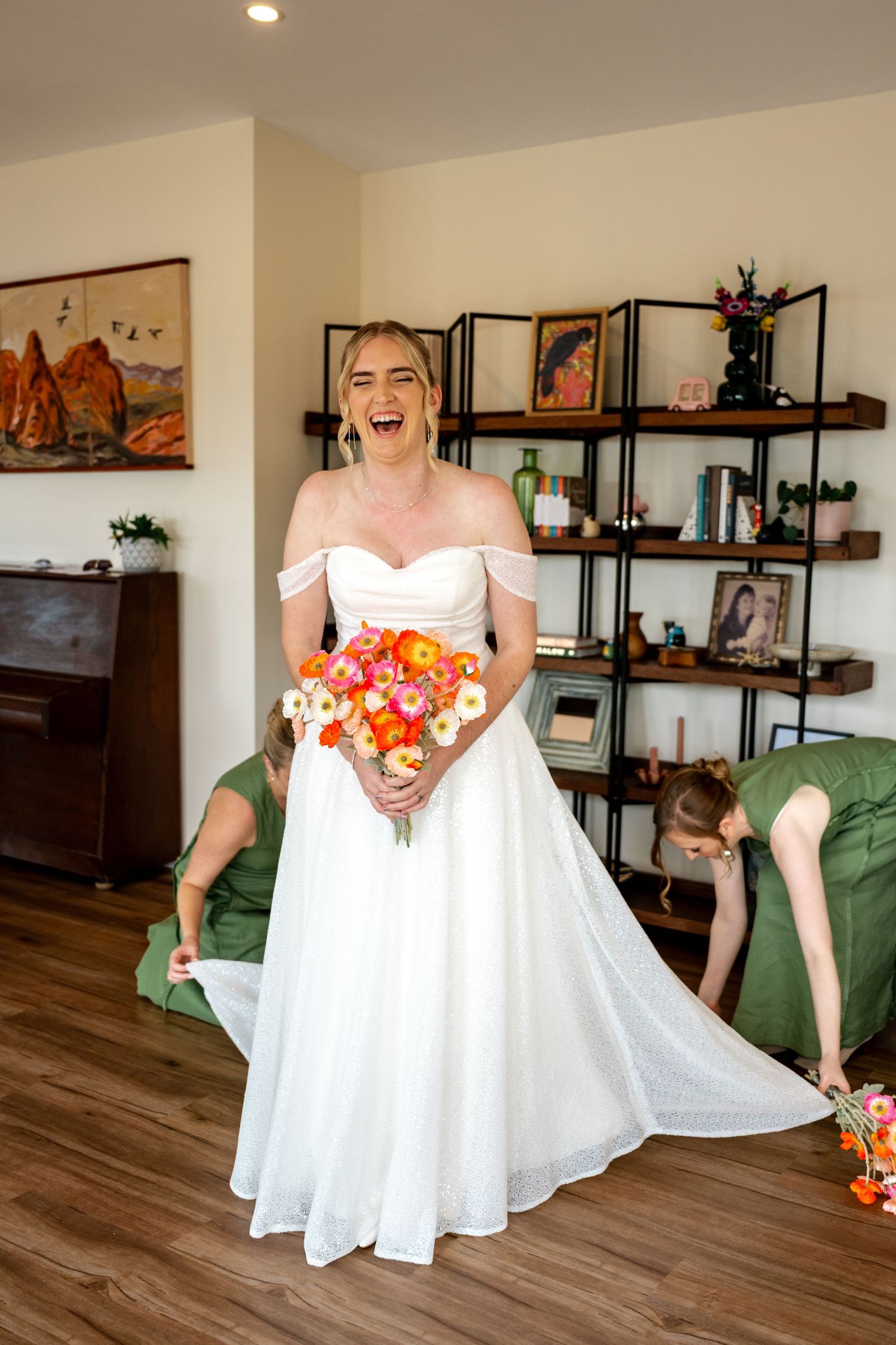 Laughing bride in white off-shoulder gown holds orange bouquet while bridesmaid adjusts her train on wooden floor.