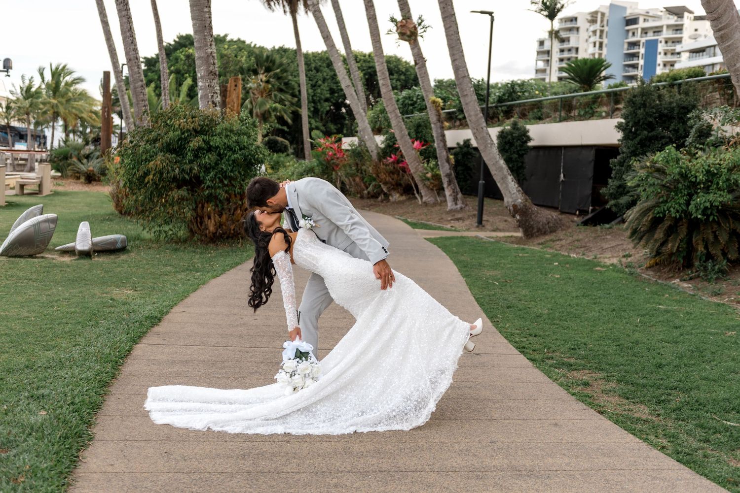 Bride and groom share a romantic dip kiss on a palm-lined garden pathway in wedding attire.