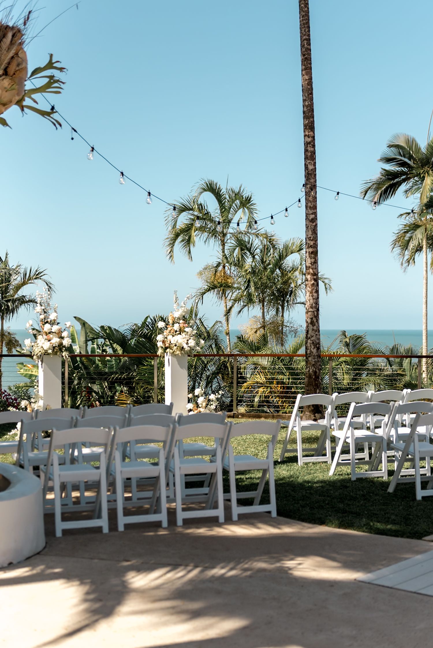 Outdoor beachside wedding ceremony setup with white chairs, floral pedestals, palm trees, and ocean views under clear sky.
