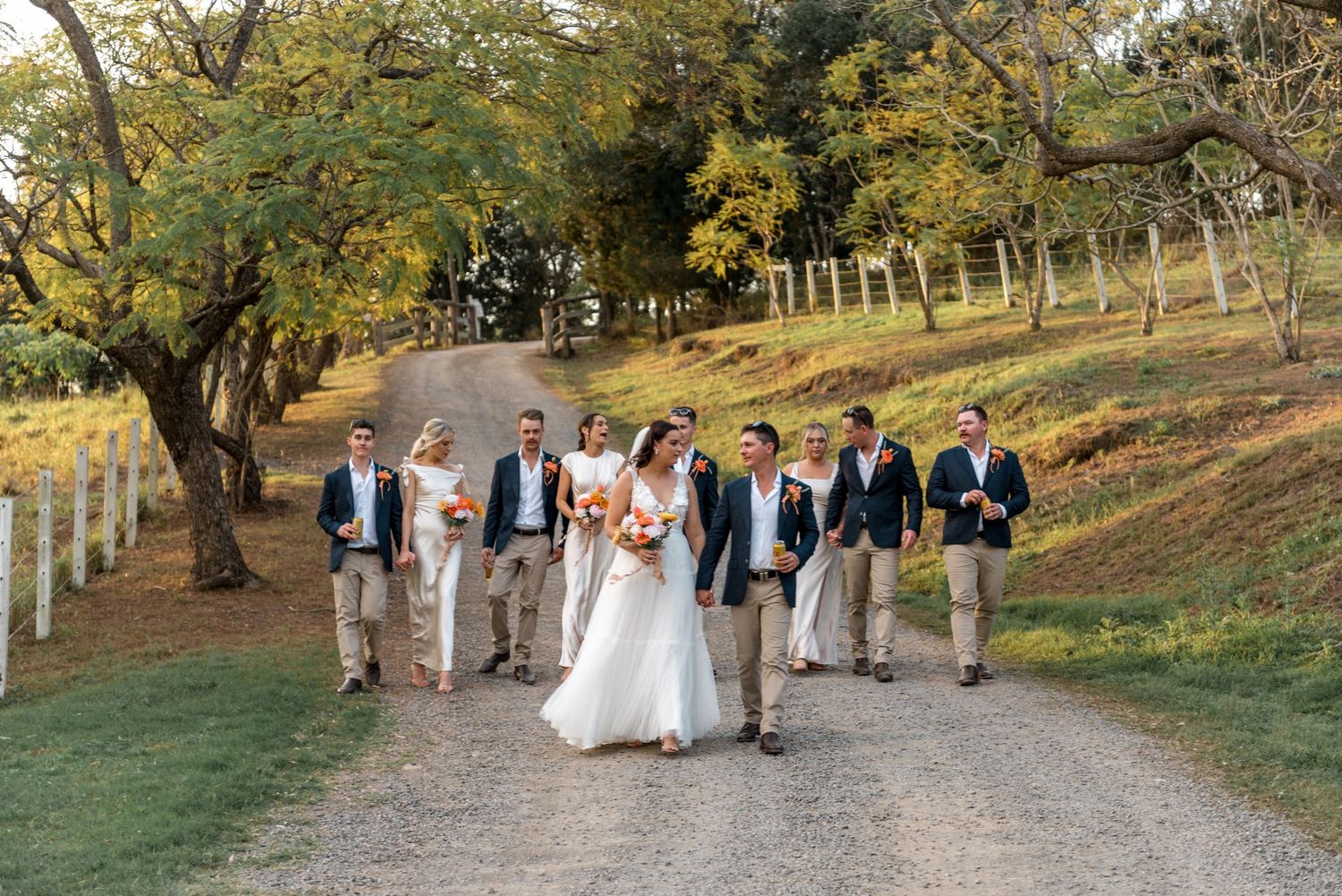 Wedding party walking along a rustic gravel driveway lined with autumn trees at sunset.