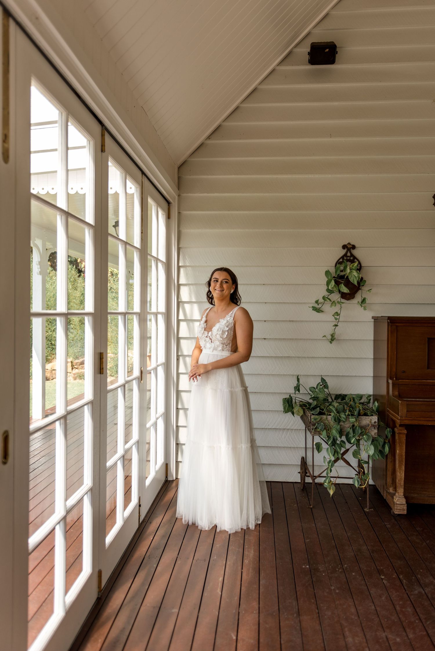 Bride in elegant white tulle wedding gown standing on sunlit timber verandah of white weatherboard home.