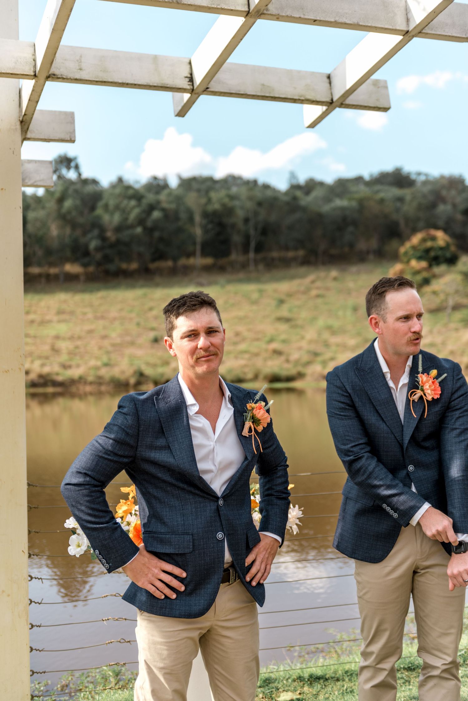 Two groomsmen in navy blazers with orange boutonnieres standing under a pergola with a scenic pond backdrop.