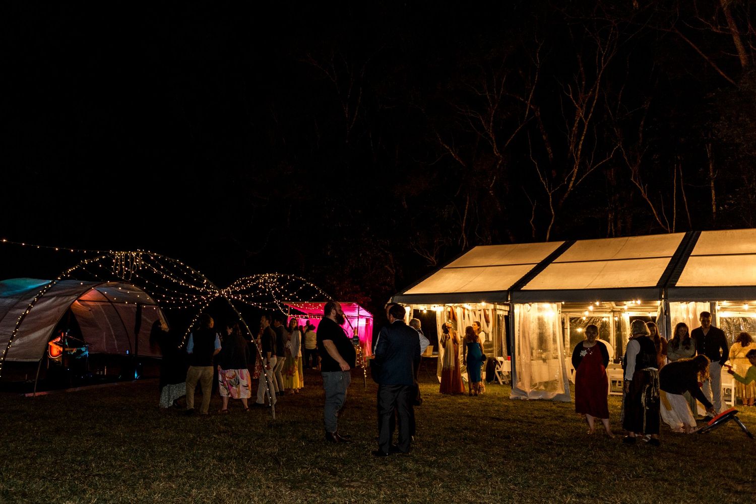 Outdoor nighttime wedding reception with string lights, a marquee tent, and guests mingling on the grass.