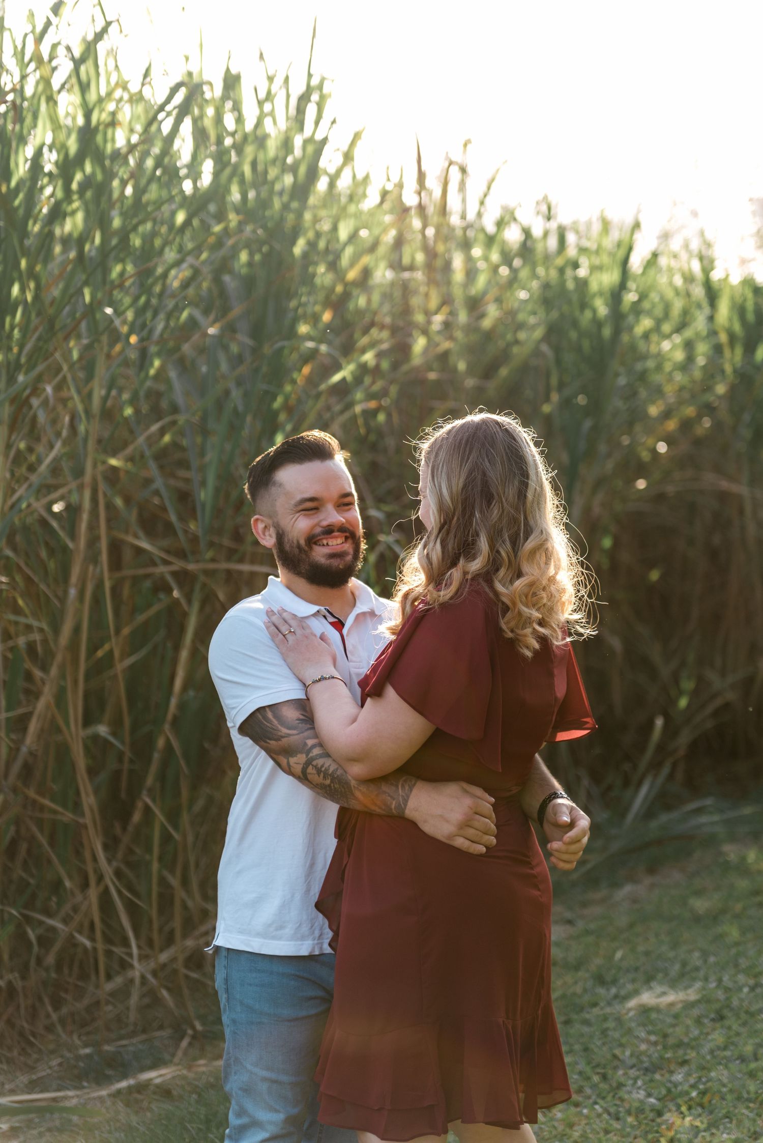 Tattooed man in white polo smiling while embracing woman in burgundy dress near tall green sugarcane field.