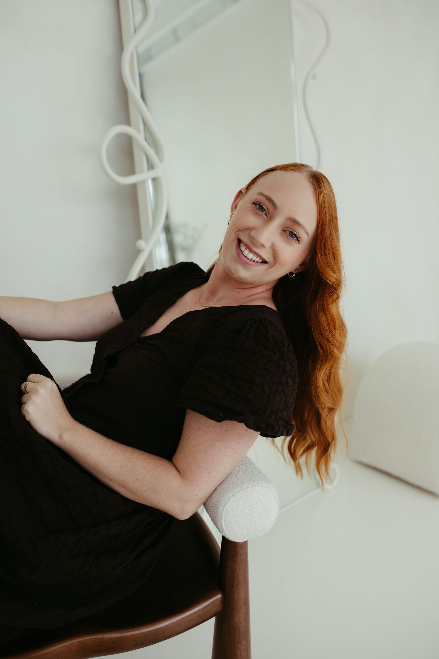 Cheerful redhead woman in black dress sitting relaxed on wooden chair, smiling.
