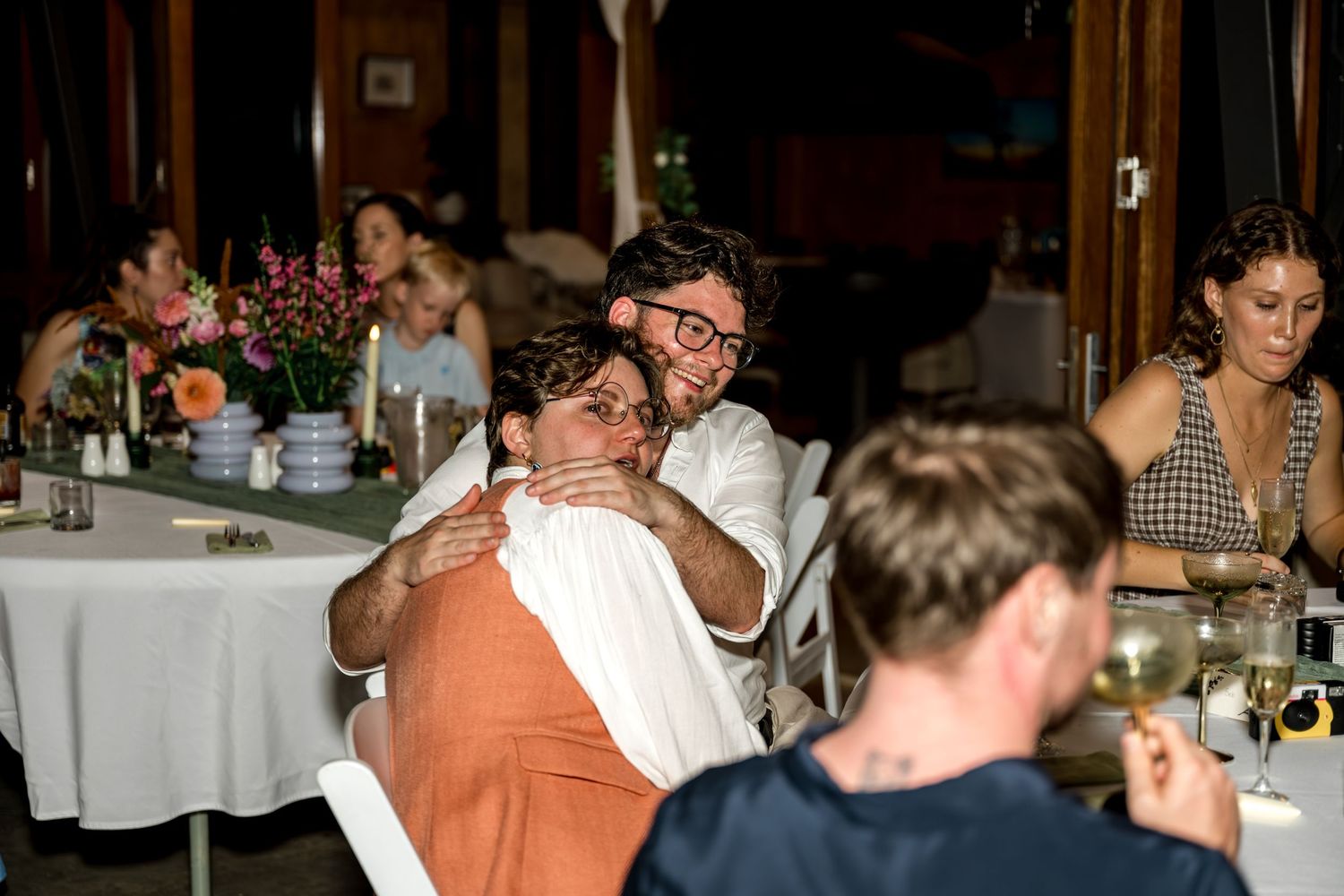 Two guests sharing a warm embrace during an indoor evening dinner reception celebration.