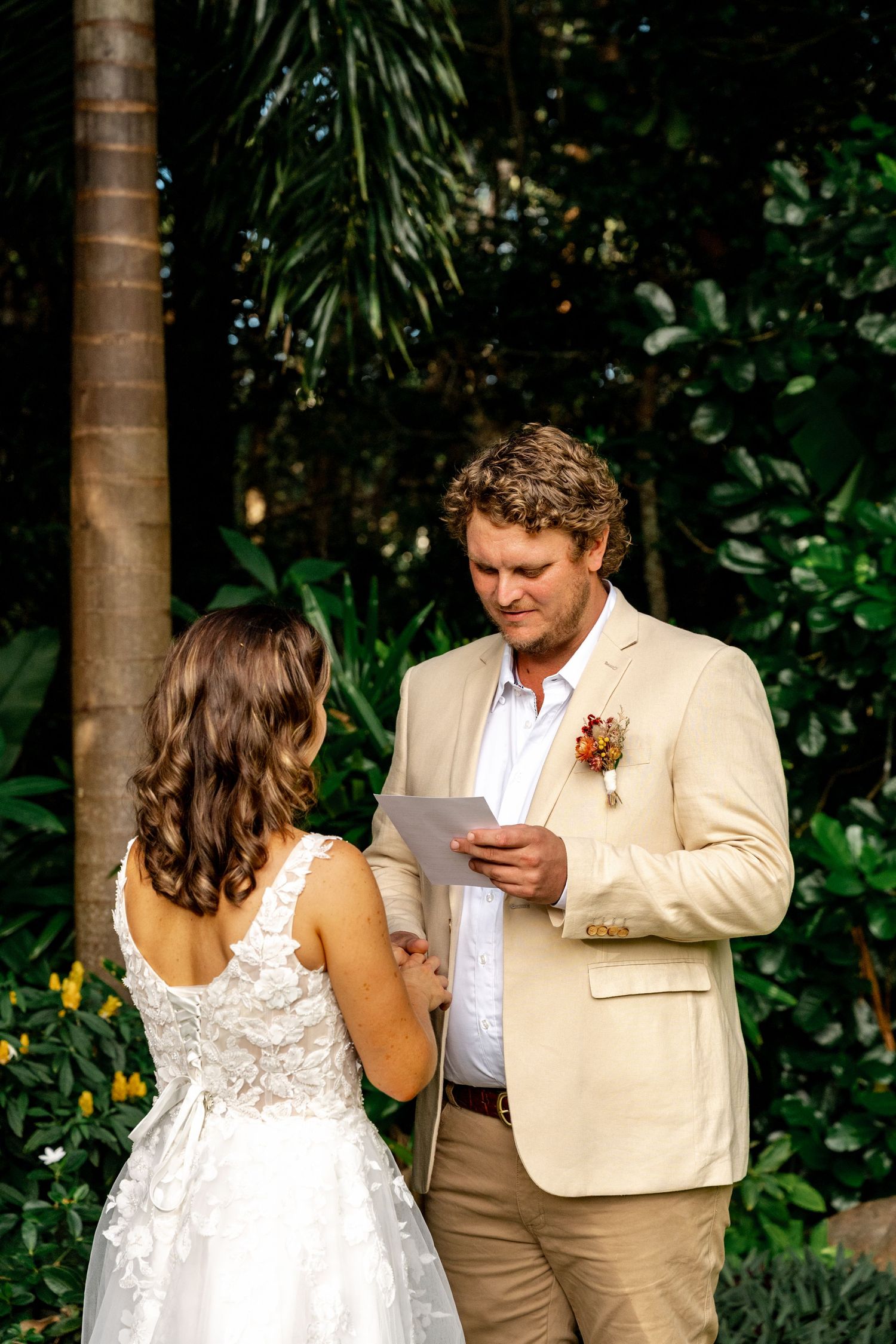 Groom in beige suit reads wedding vows to bride in white lace dress during outdoor garden ceremony.