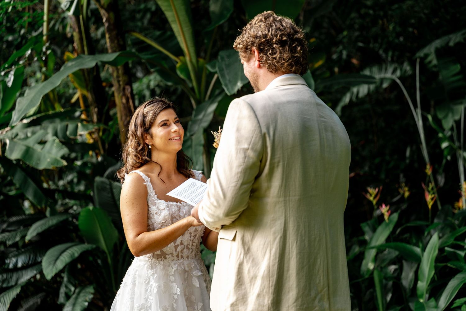 Bride gazes at groom while exchanging vows during lush tropical garden wedding ceremony.
