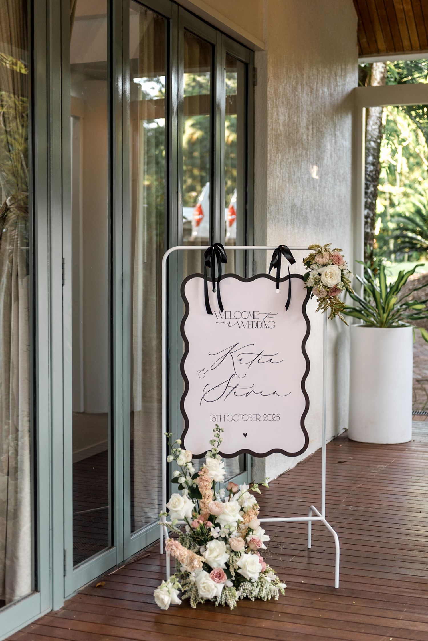 Elegant wedding welcome sign for Katie and Simon on white stand with pink floral arrangements on timber porch.