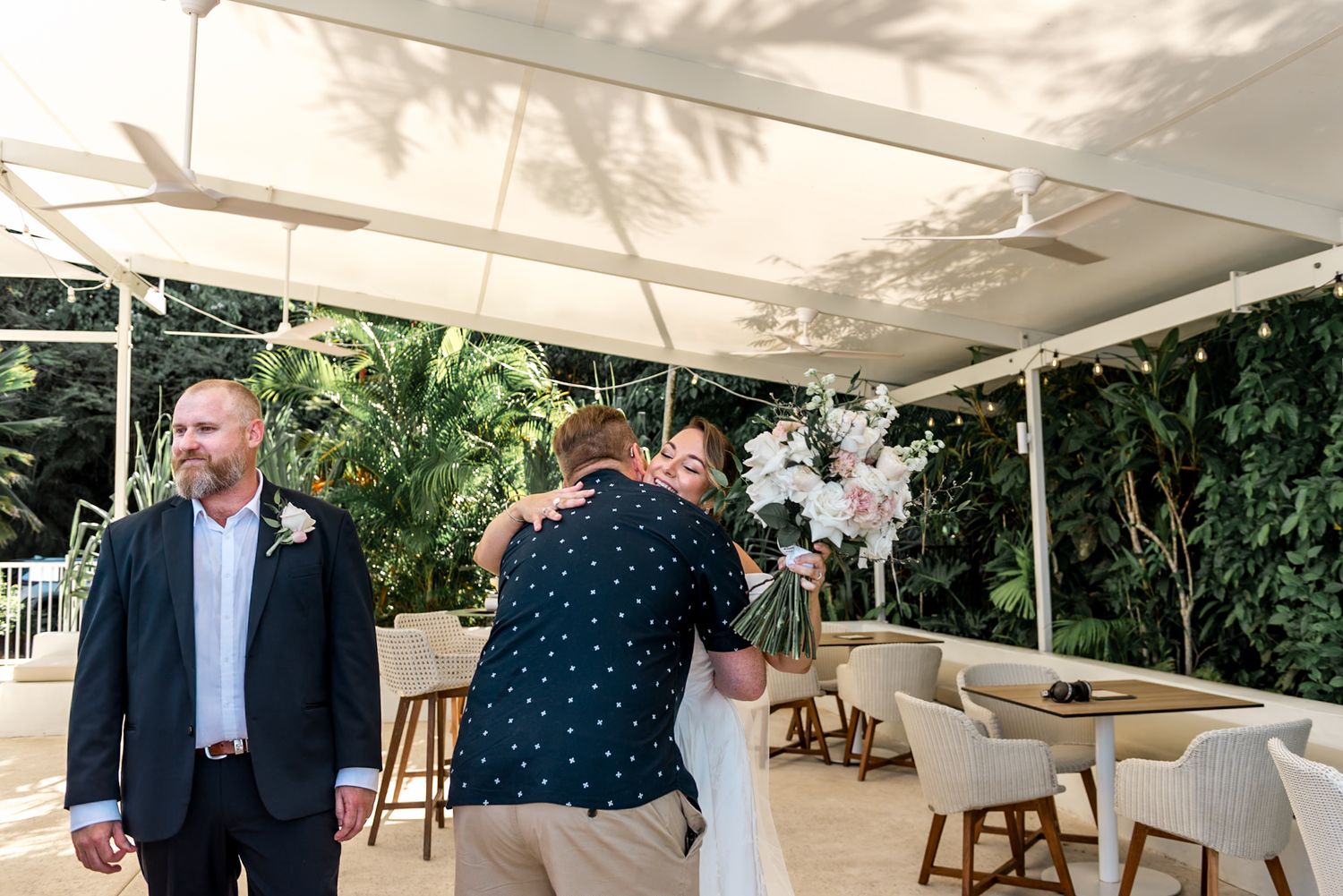 Bride receives congratulatory hug from guest while groom stands nearby at outdoor wedding.