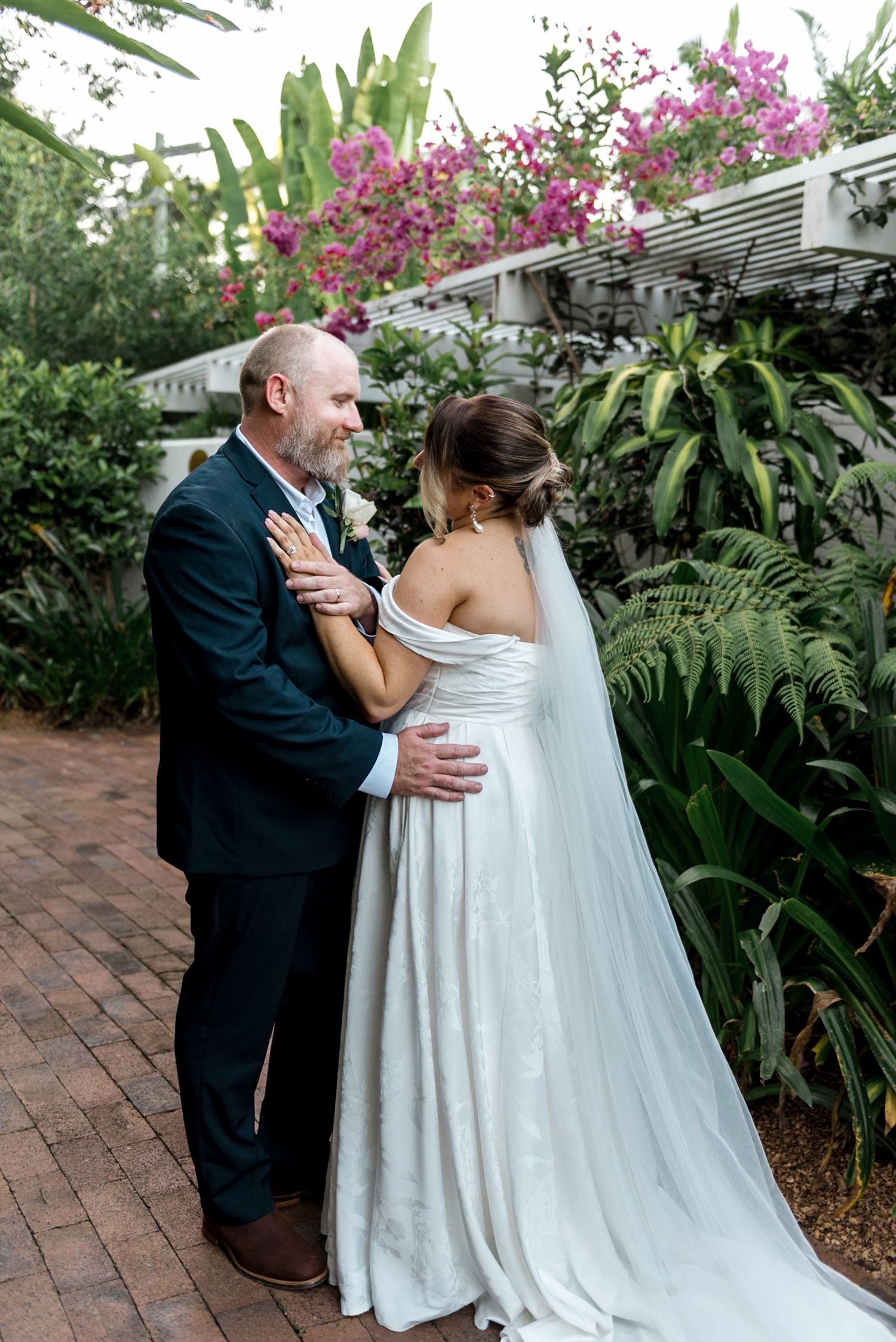 Bride and groom sharing tender moment in tropical garden with pink bougainvillea blooms.