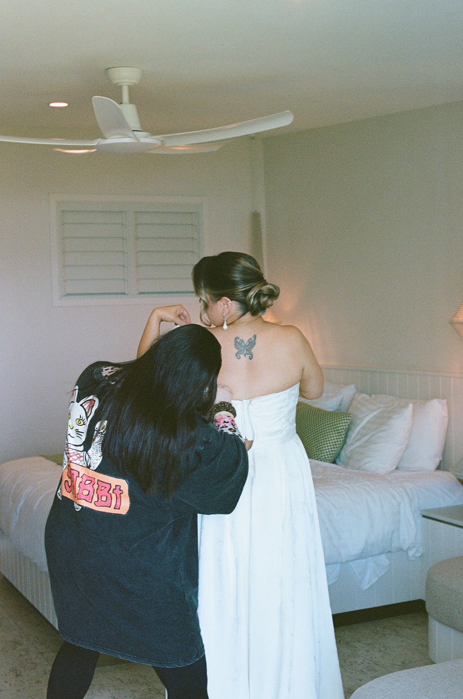 Bridesmaid in black jacket helps bride with white wedding dress in bright hotel room.