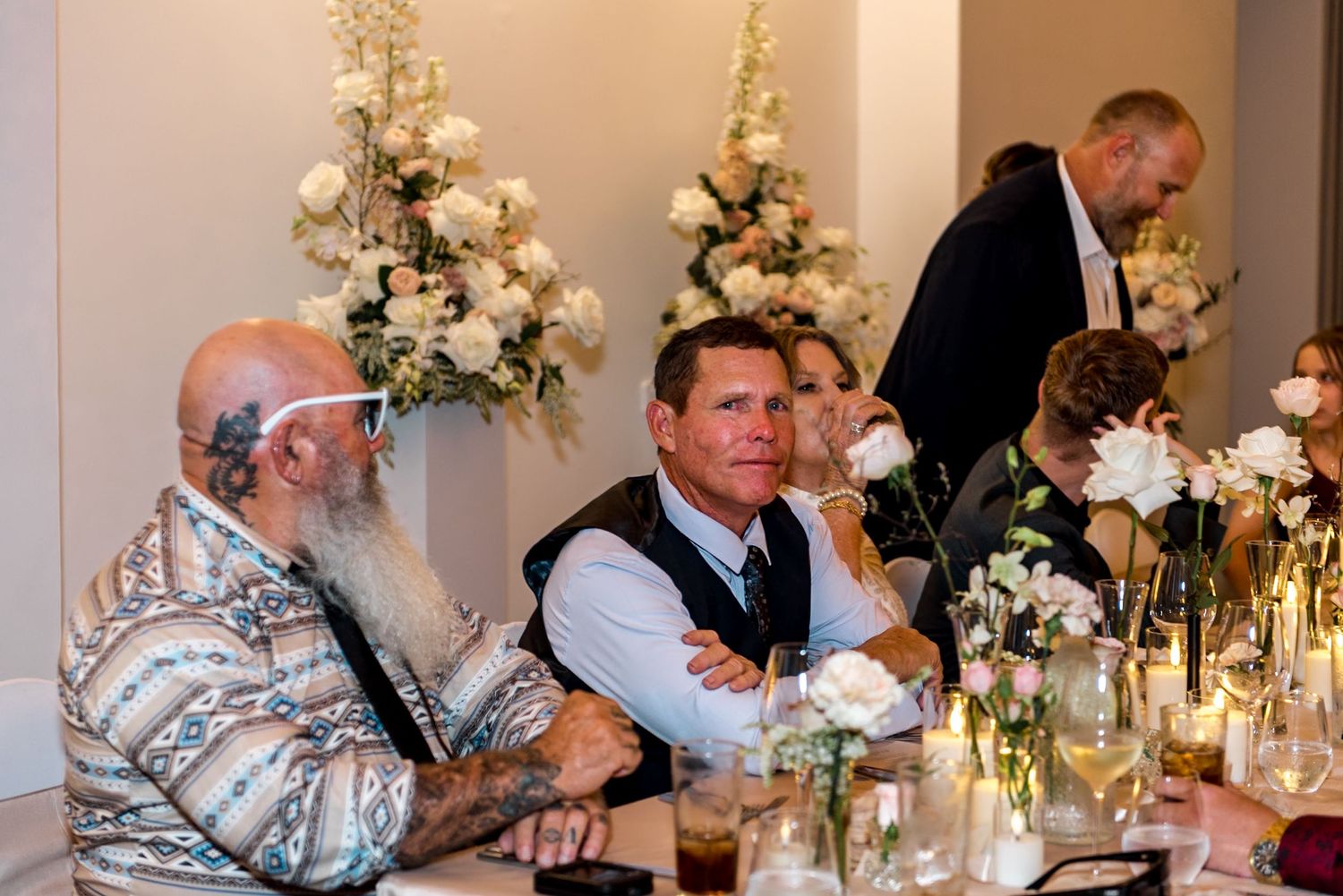 Two male wedding guests chat at elegantly decorated reception table with white floral centerpieces.