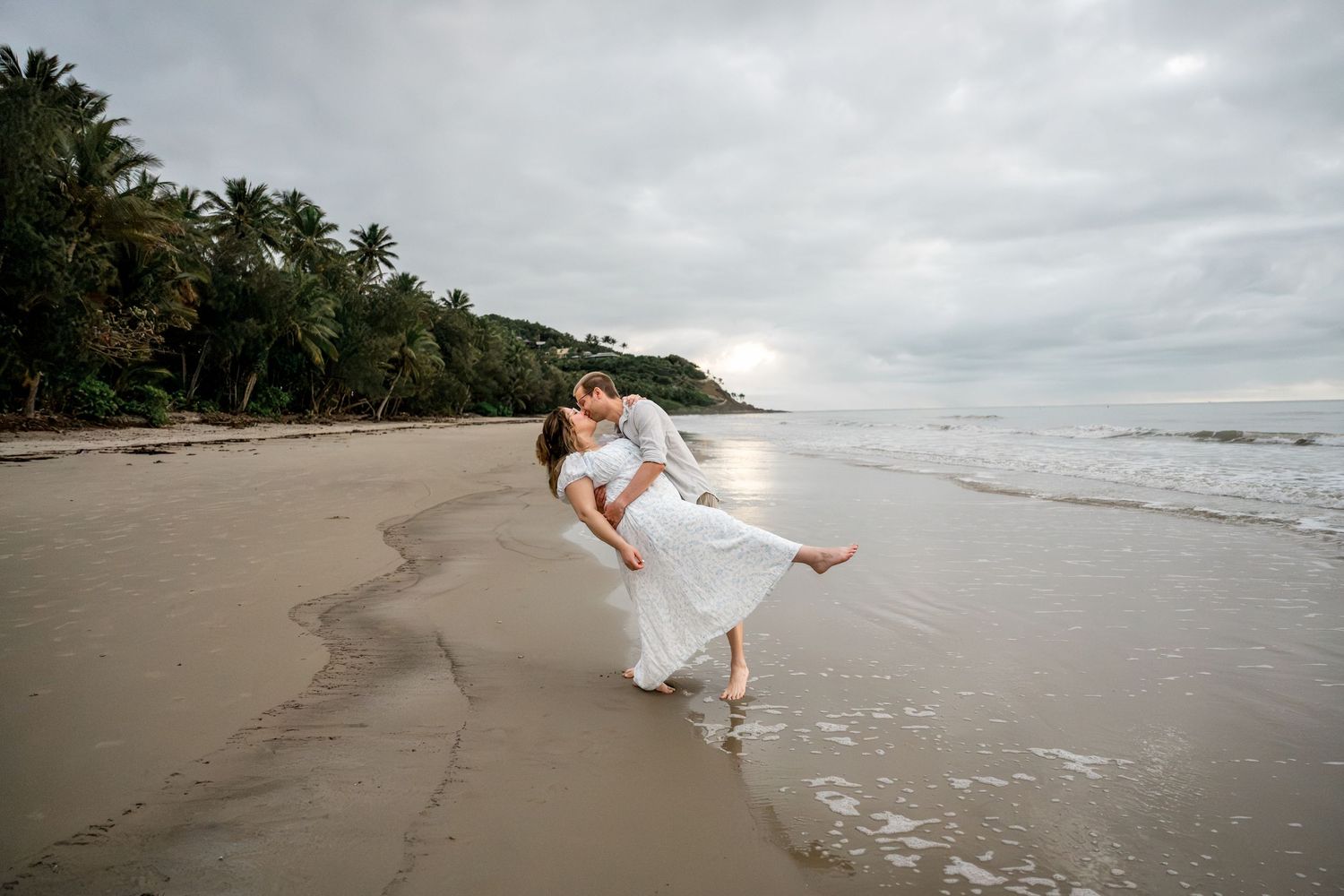 Man lifting woman in white dress for a romantic dip on a tropical sandy beach at Four Mile Beach, Port Douglas.