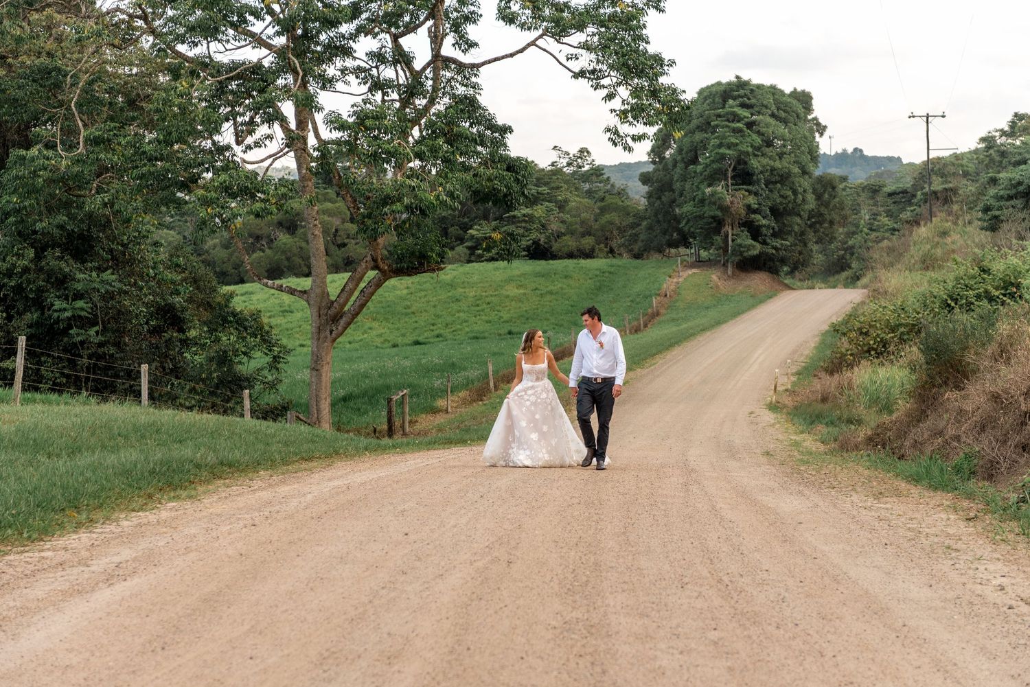Bride and groom walking hand in hand down a scenic dirt road surrounded by lush green fields.