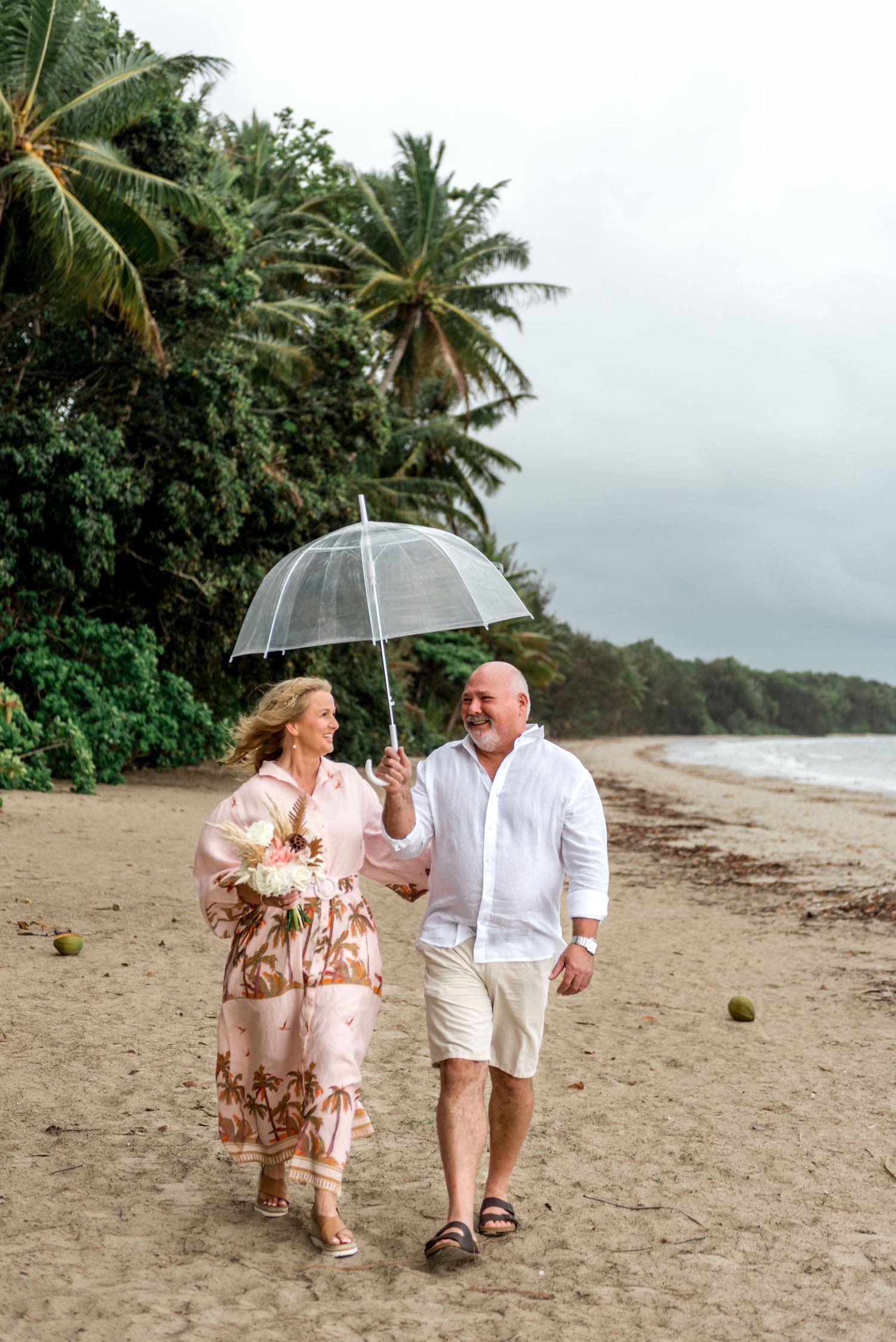 Happy couple walking on a tropical beach under a clear umbrella surrounded by palm trees.