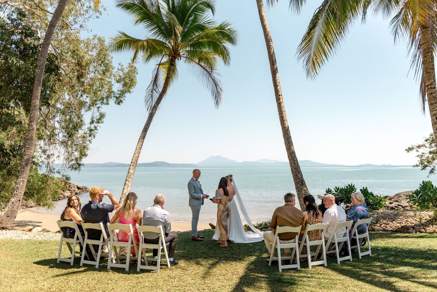 Sunny tropical beach wedding ceremony with guests in white chairs framed by tall swaying palm trees.