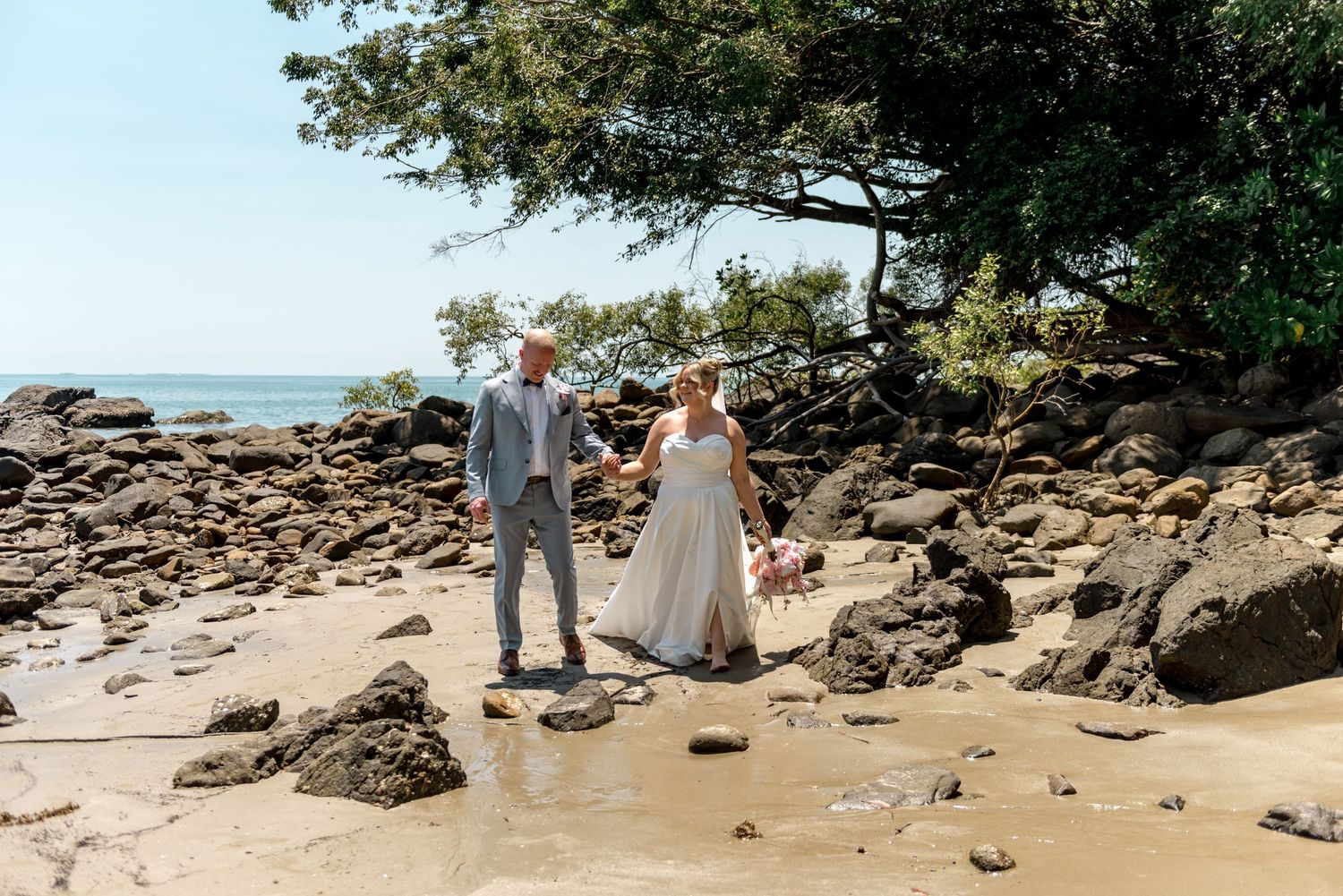 Newlyweds holding hands on a scenic rocky beach with lush green trees and ocean views.