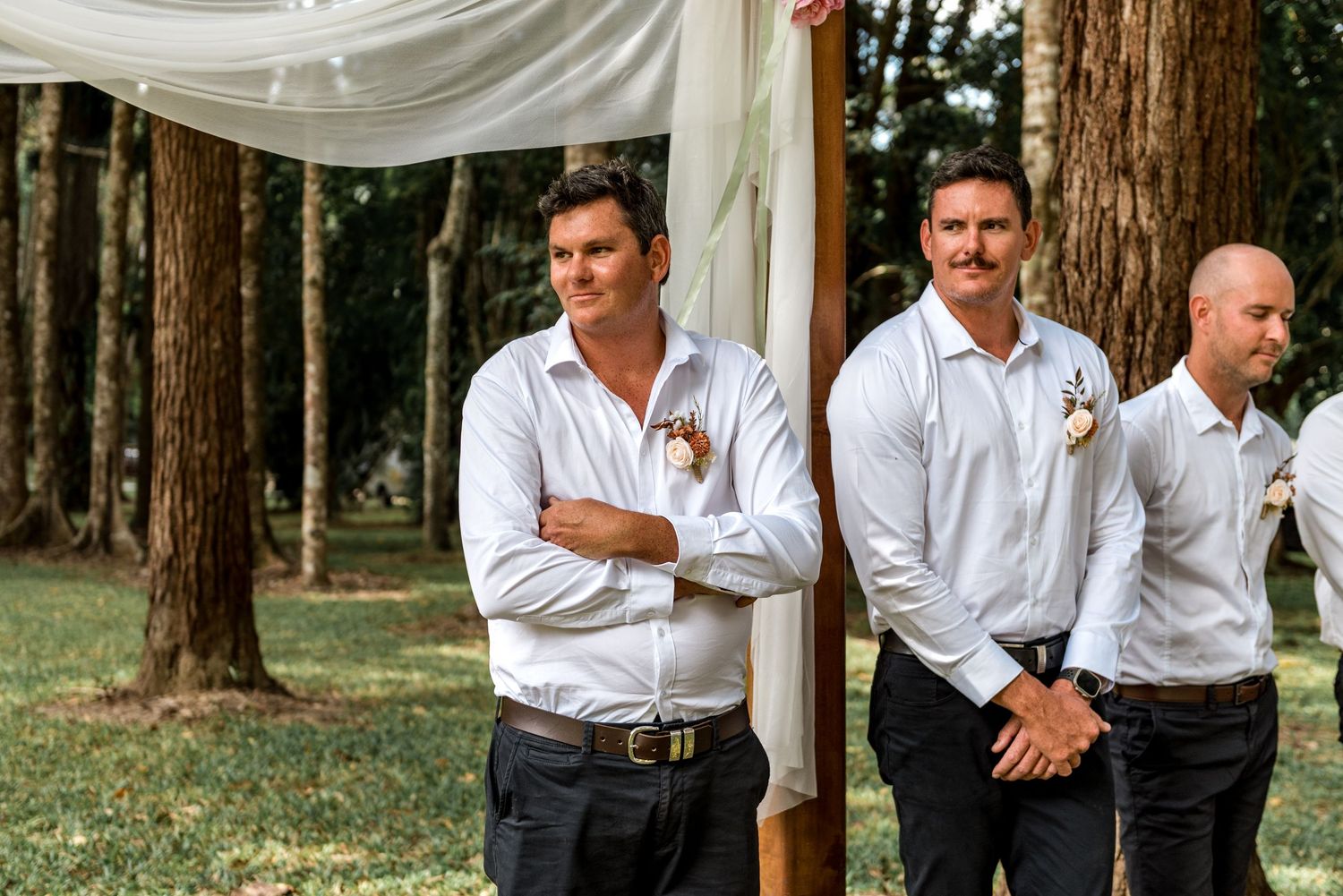 Three groomsmen in white shirts stand attentively at an outdoor woodland wedding ceremony.