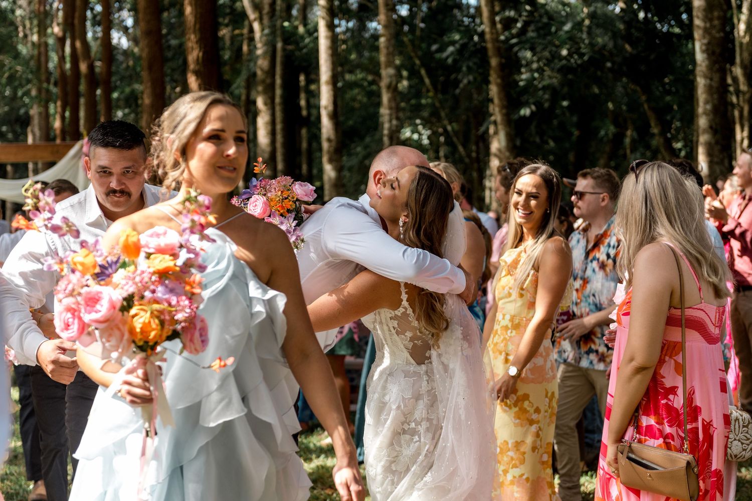 Bride in lace gown holds colorful bouquet while receiving warm hugs from guests at outdoor forest wedding.