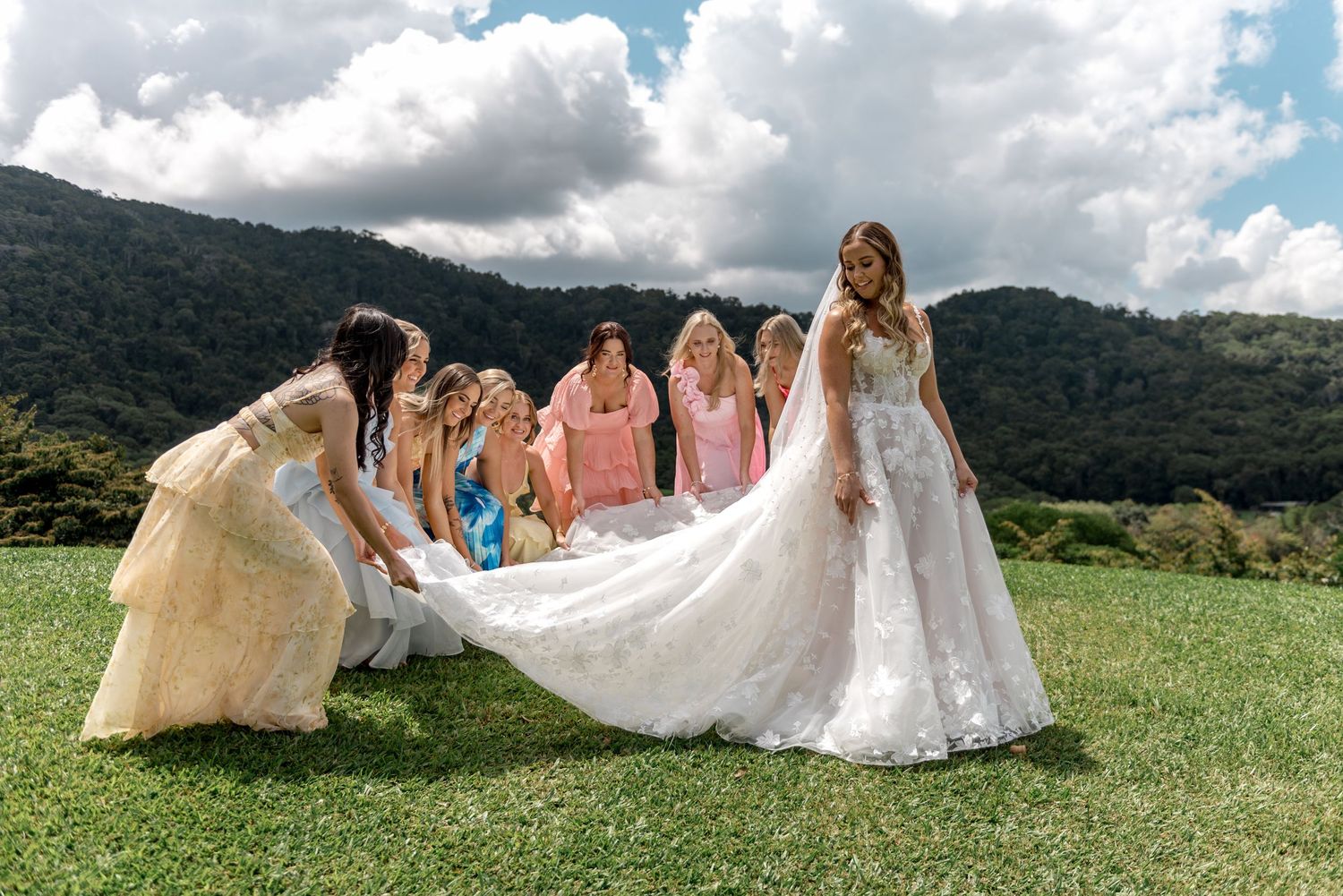 Bridesmaids in colorful dresses help arrange bride's long lace train on green lawn outdoors.
