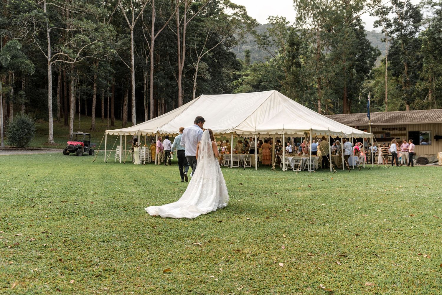 Bride and groom share a romantic moment on green lawn beside a white wedding reception marquee tent.