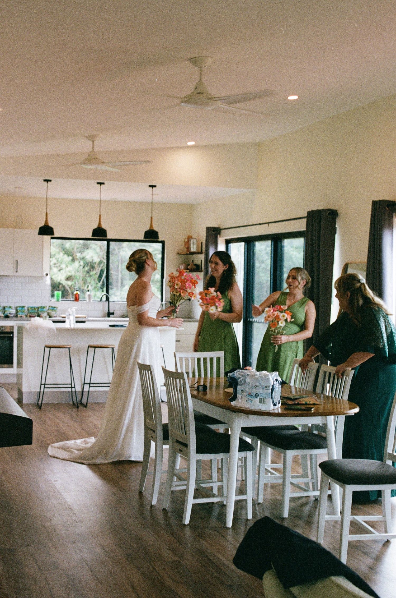 Bride in white gown with bridesmaids in green dresses preparing for wedding in a modern kitchen.