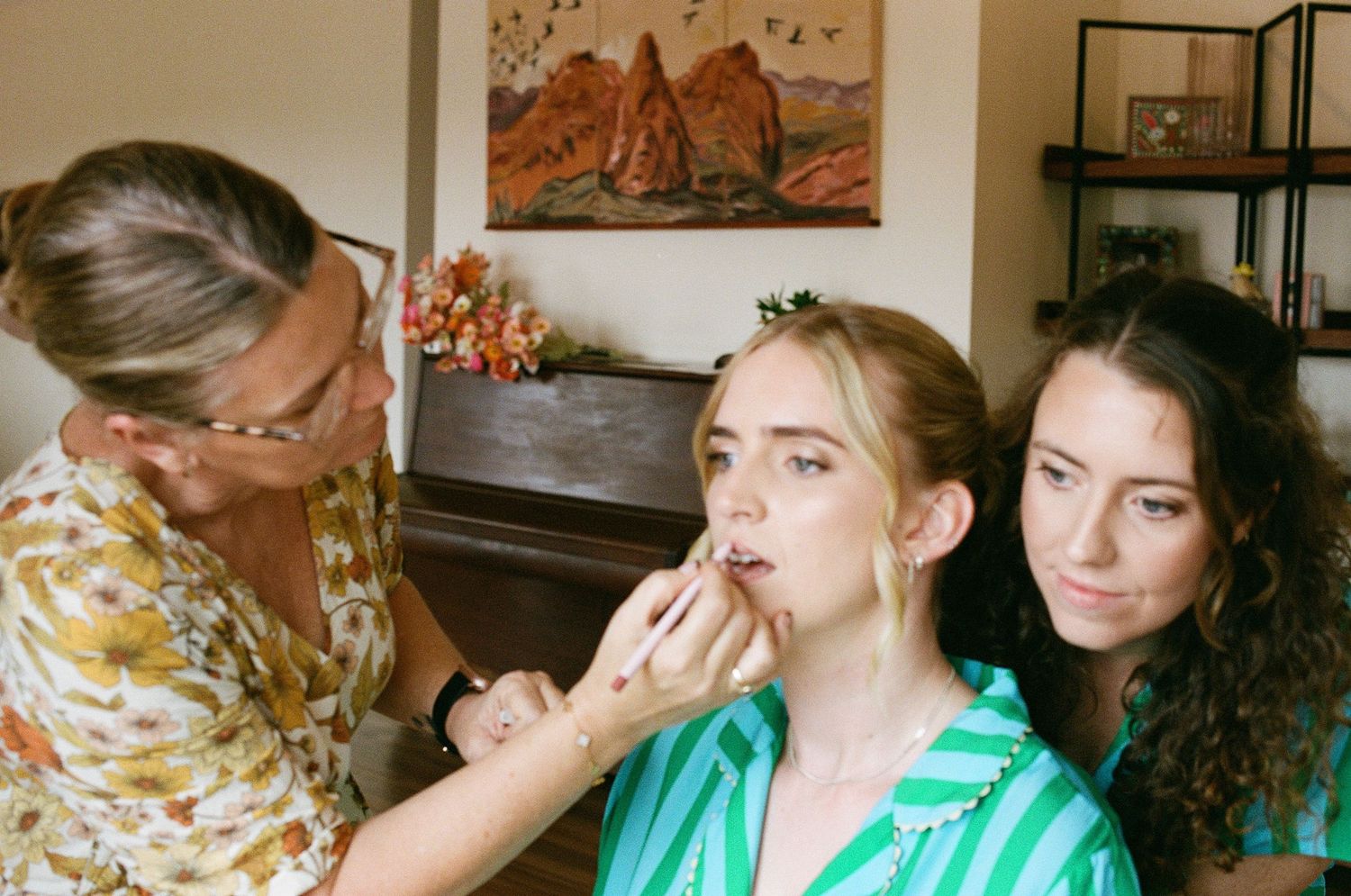 A makeup artist applies lipstick to a bride wearing a floral crown during wedding prep.