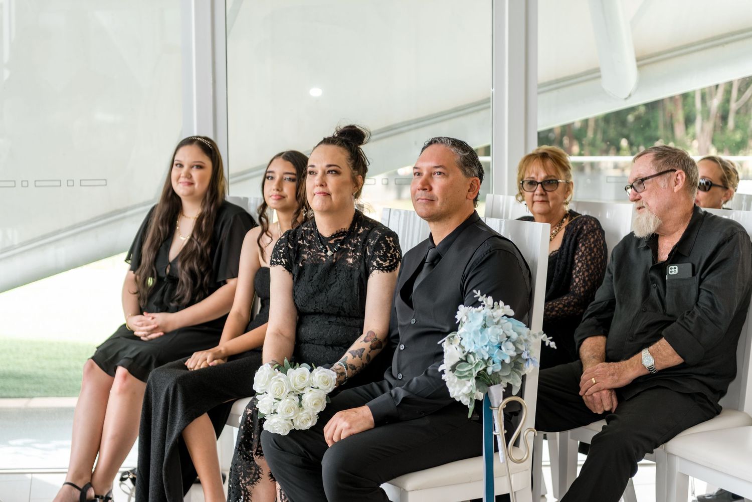 Wedding guests dressed in black attire seated at ceremony, holding light blue floral bouquets.
