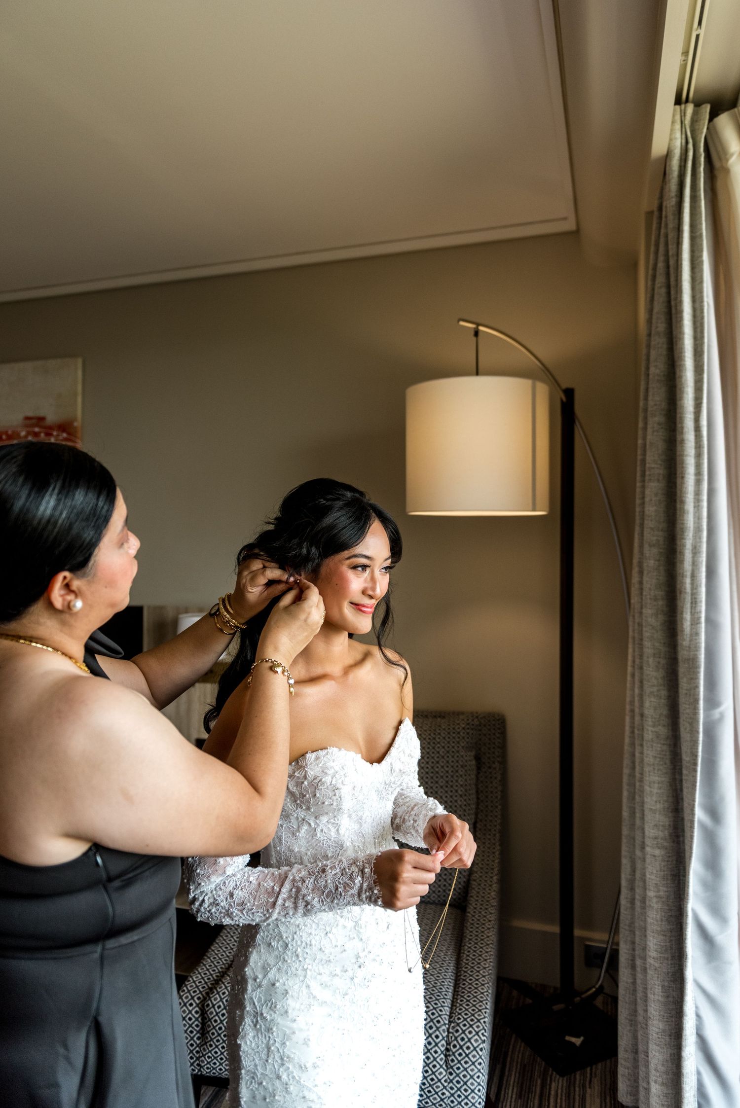 Mother helping smiling bride put on earrings while dressed in white lace wedding gown.
