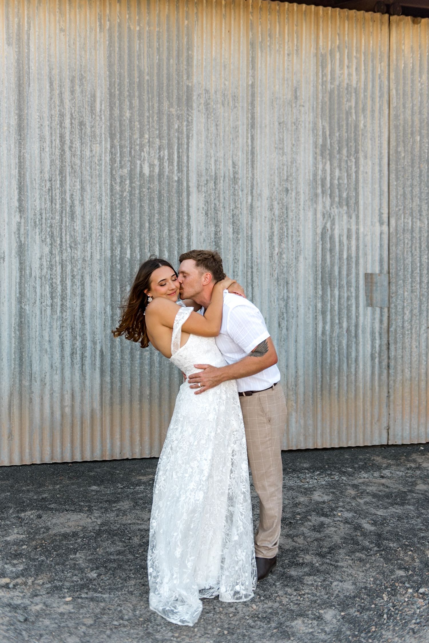 Groom kisses bride on cheek as she smiles joyfully in lace gown before corrugated metal backdrop.