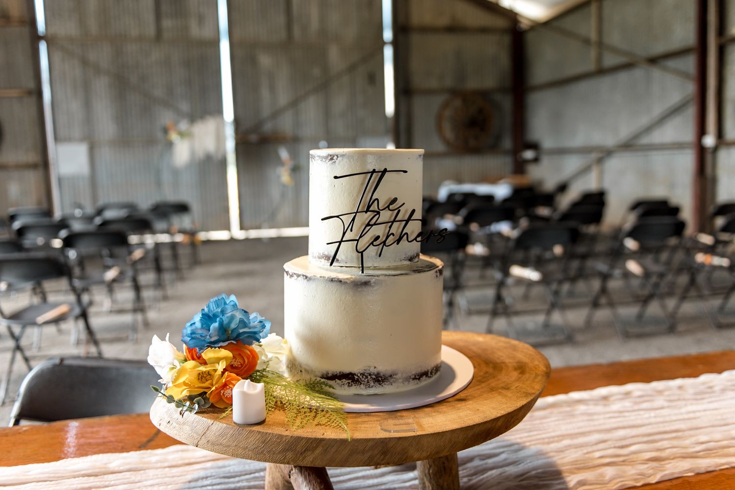 Two-tier semi-naked Fletcher wedding cake displayed on a rustic wooden stand in a barn venue.