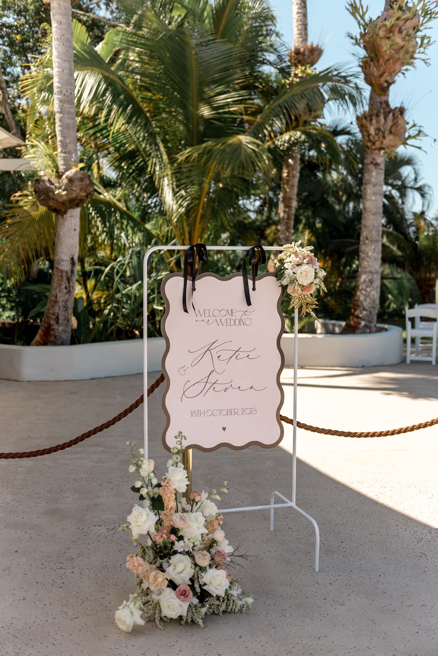 Elegant wedding welcome sign for Kate and Steve displayed on a white stand with blush floral arrangements and palm trees.