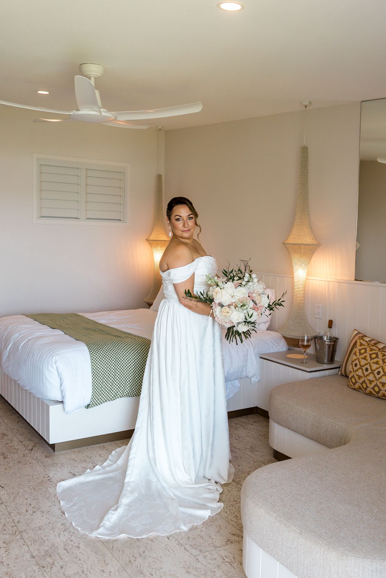 Bride in off-shoulder white gown holding white floral bouquet in a warmly lit hotel room.