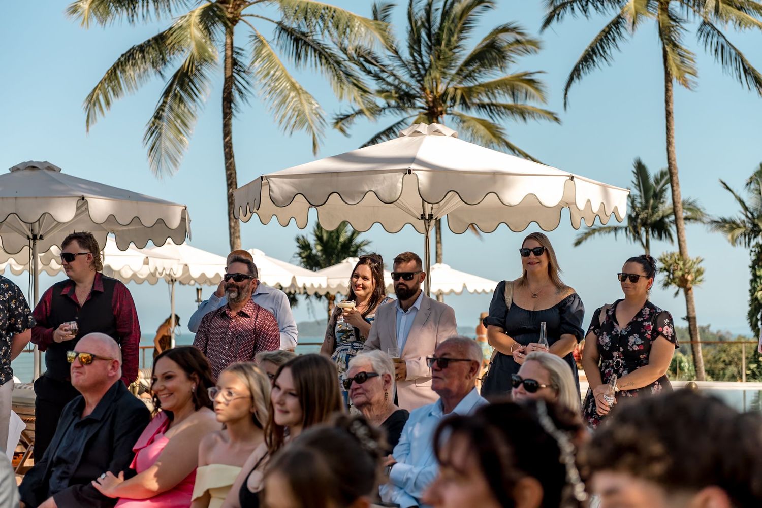 Guests gathered outdoors at a tropical wedding ceremony with white umbrella shade structures and palm trees.