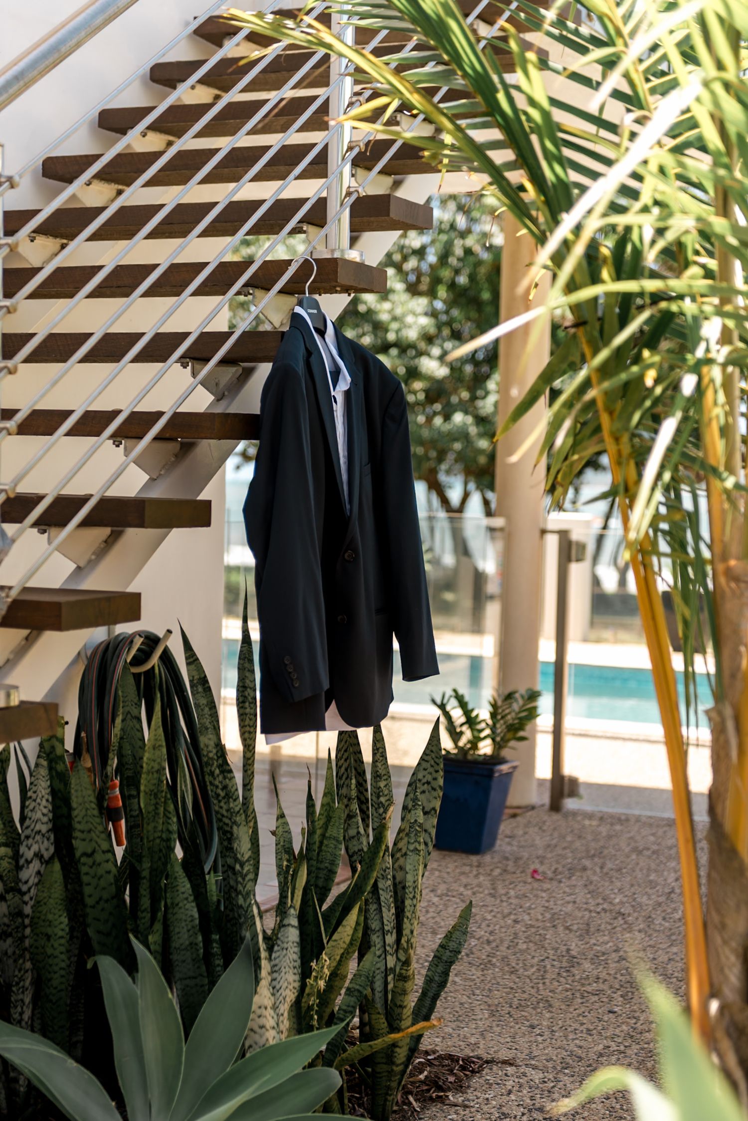 Dark navy groom suit hanging on wooden staircase railing surrounded by tropical plants near pool.