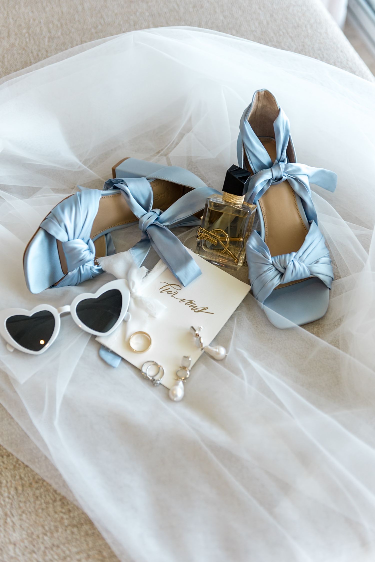 Bridal accessories flat lay with powder blue knotted heels, heart sunglasses, pearl earrings, and perfume.