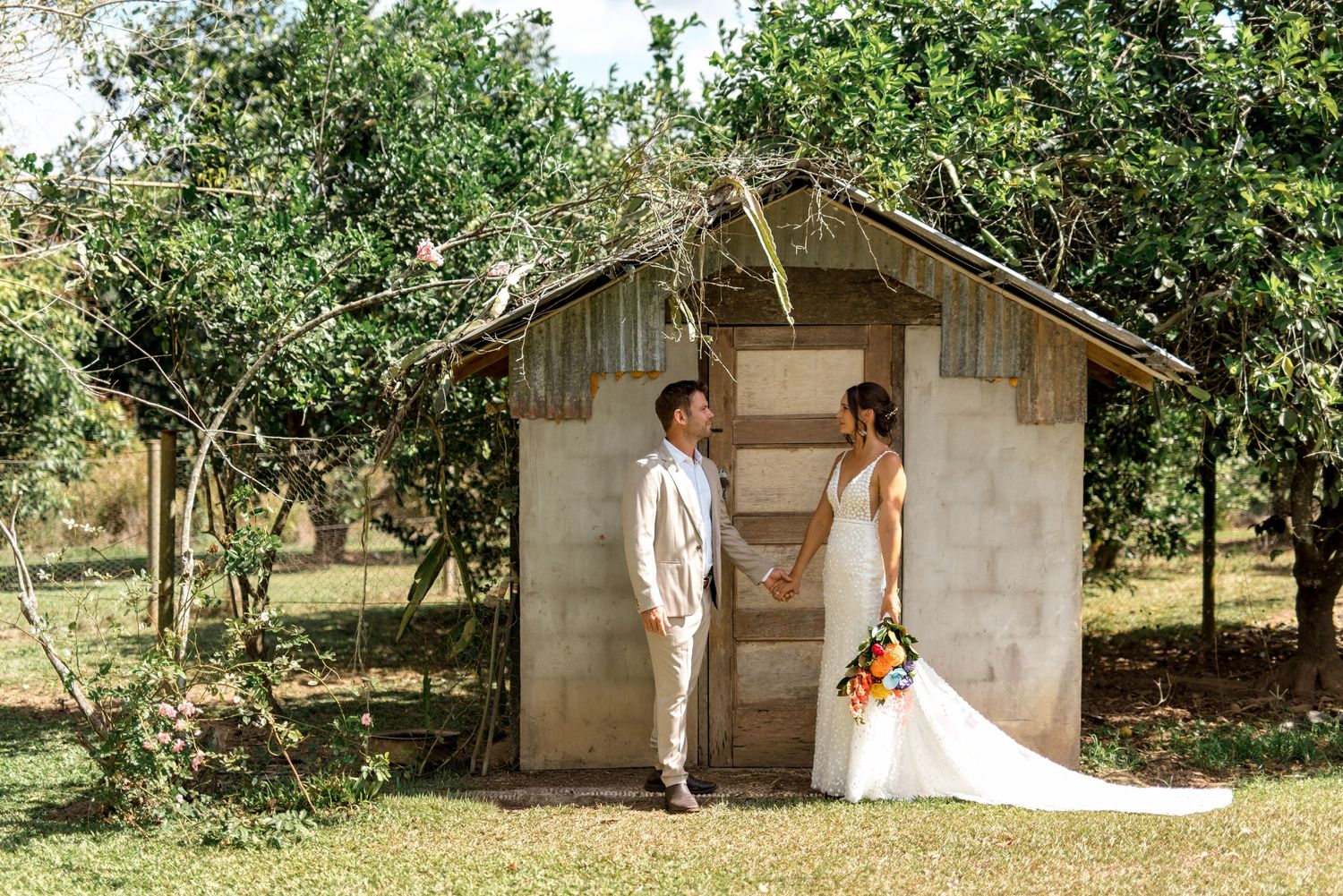 Bride and groom hold hands beside a rustic stone shed surrounded by tropical greenery.
