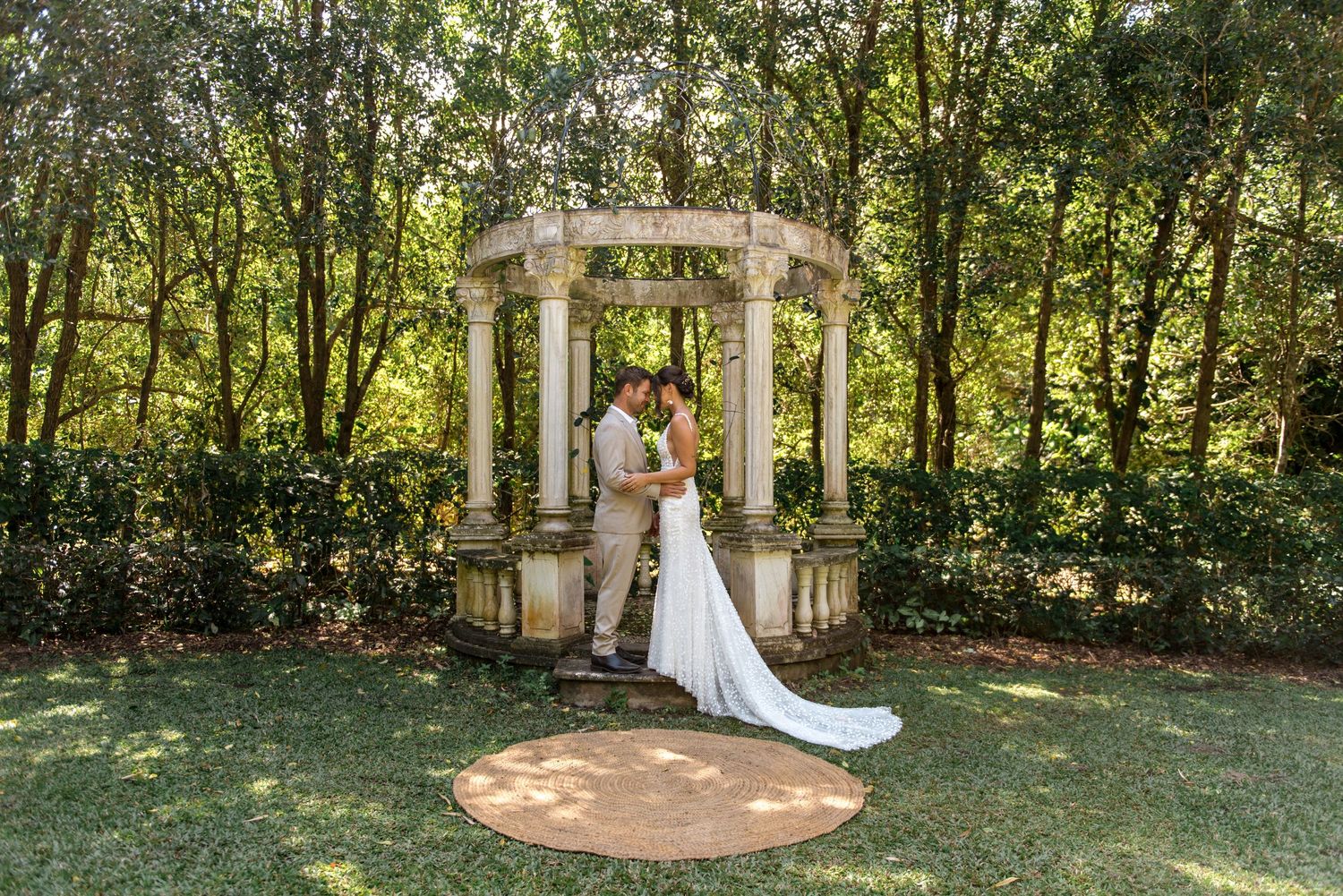 Bride and groom kissing under a classic stone columned gazebo surrounded by lush green garden foliage.