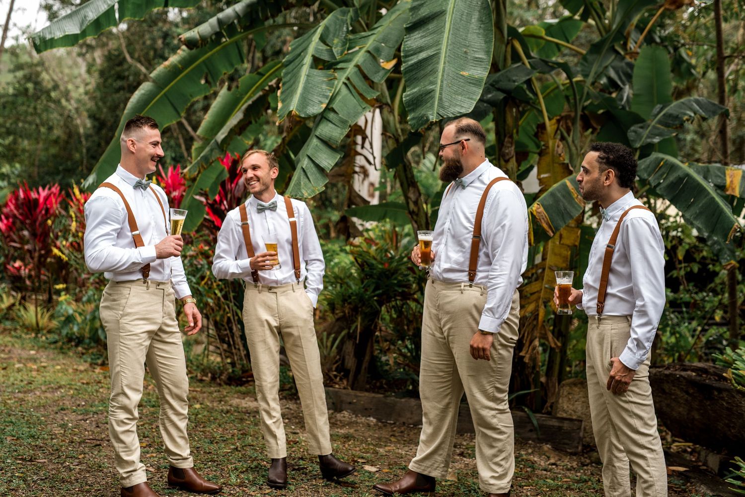 Groomsmen in khaki pants and suspenders laughing together outdoors among tropical plants.