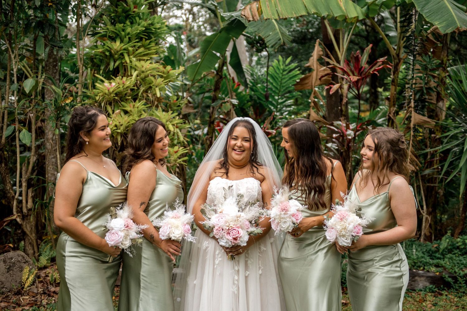 Smiling bride in white gown laughs with bridesmaids in sage green dresses holding pink floral bouquets.