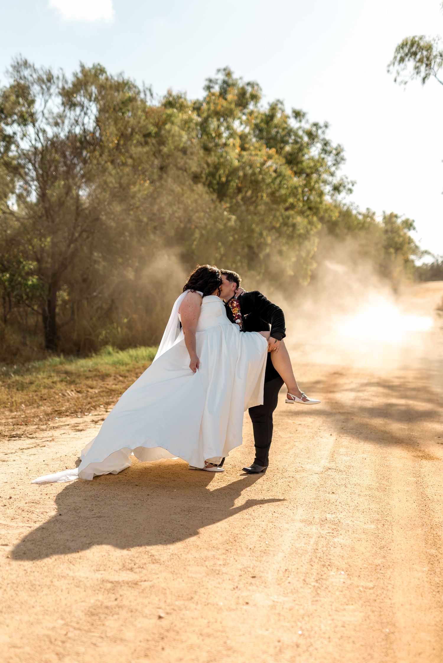 Same-sex couple sharing a romantic dip kiss on a dusty outback dirt road, bride in white gown and partner in black suit.