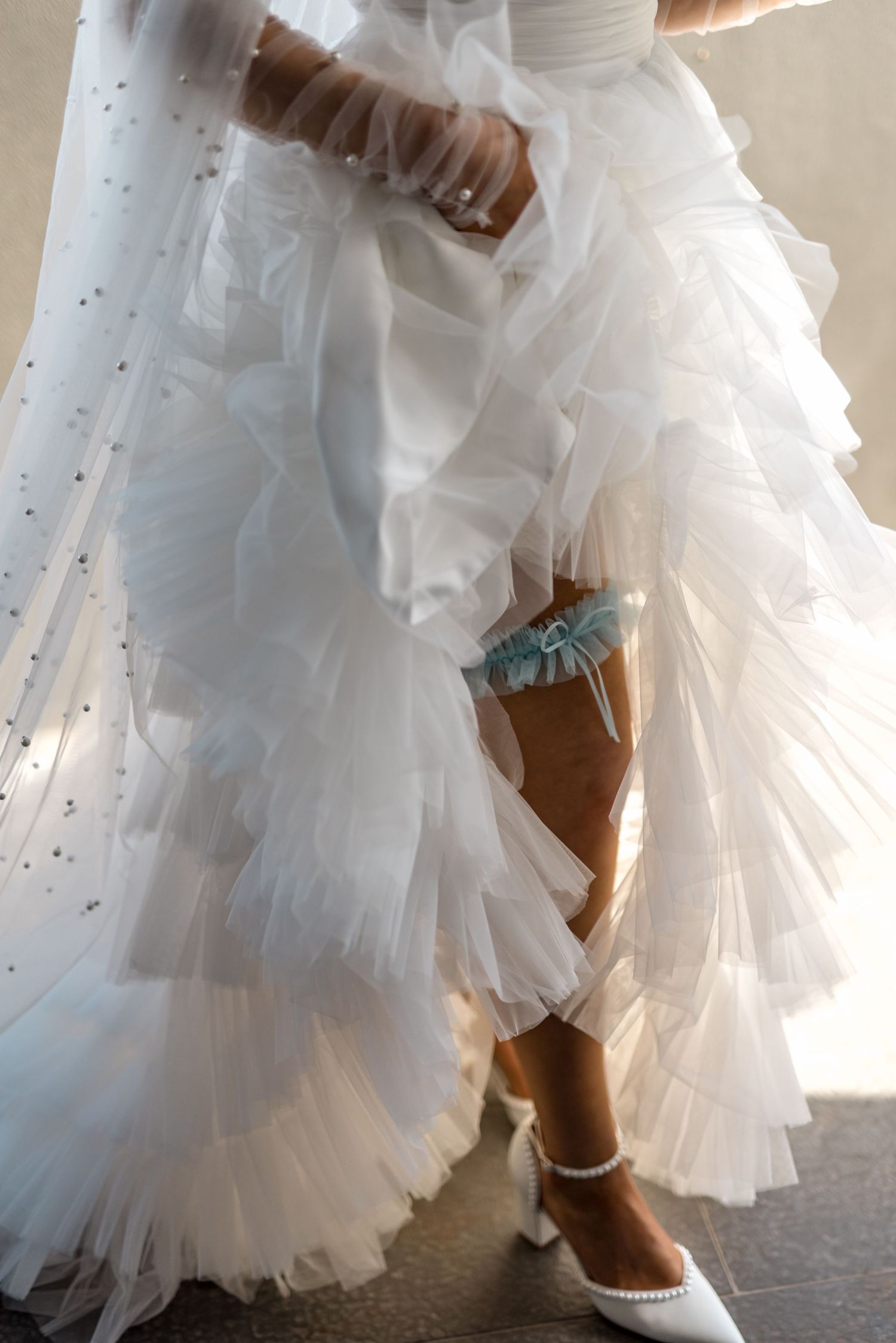 Bride in layered tulle gown displaying blue garter and pearl wedding heels.