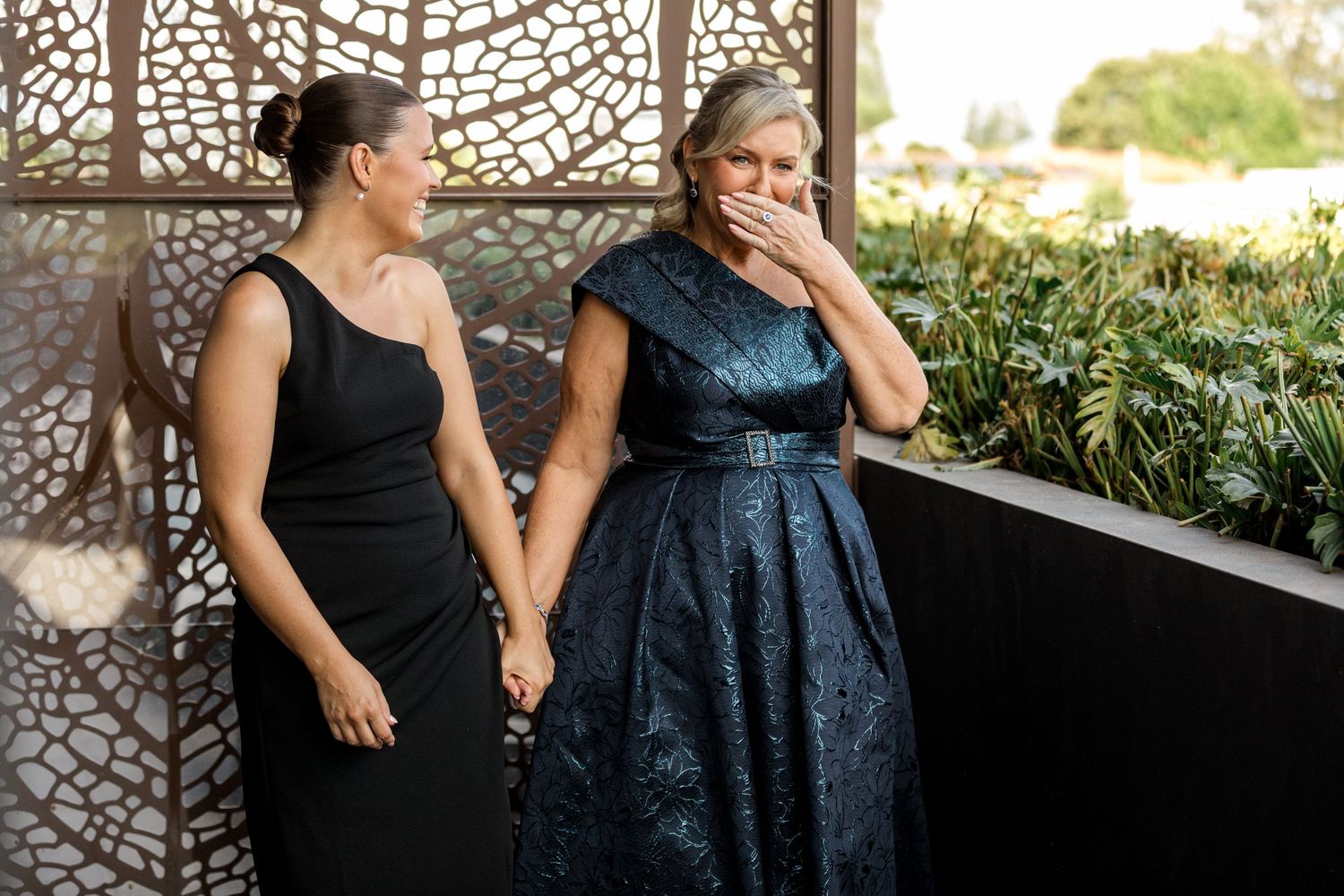 Two women in formal attire holding hands and laughing emotionally at an outdoor wedding venue.