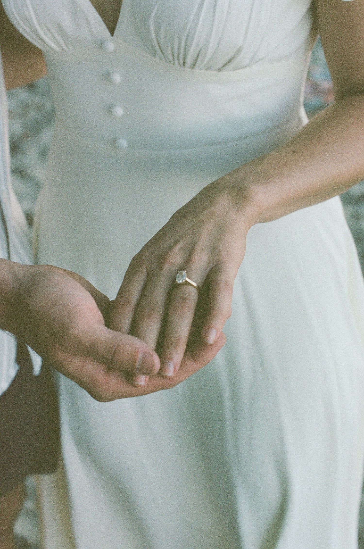 Close-up of engagement ring on womans hand in white dress held by partner on beach.